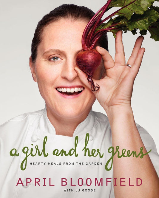 A woman in a chef's uniform smiles while holding up a fresh beet by its greens beside her head, against a plain background. The text reads "A Girl and Her Greens: Hearty Meals from the Garden by April Bloomfield (Author), JJ Goode EdD. (Author)